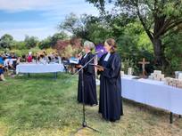 Pfarrerin Schlarb und Frau Schücking-Jungblut im Talar halten eine Ansprache an die Gemeinde beim Tauffest, dahinter Altartisch mit weißem Tischtuch, darauf Schatzkästchen mit den Namen der Täuflinge. Im Hintergrund der Posaunenchor.